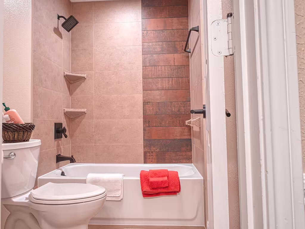 Upgraded bathtub remodel with beige wall tile, rustic wood-look accent panel, matte black fixtures, and corner shelves
